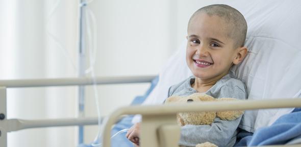 A little boy with a shaved head is smiling as he is laying on a hospital bed. He is battling with cancer.